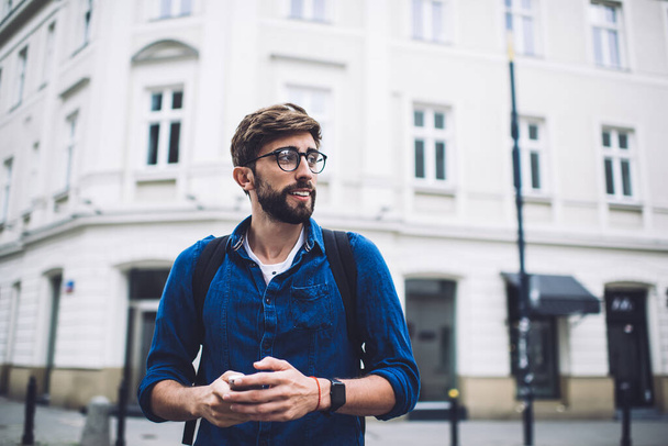 From below of positive young hipster male in casual clothes with smartphone and backpack wearing eyeglasses in city street on daytime looking away - Foto, Imagem