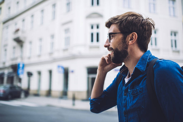 Side view of young bearded male in casual clothes and eyeglasses talking on mobile phone while walking in city and looking away - Valokuva, kuva