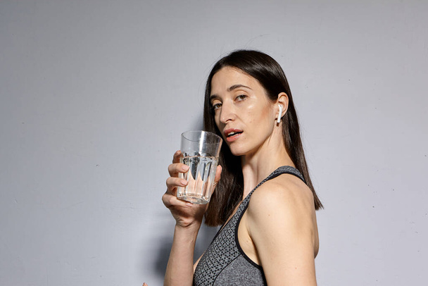 Женщина в спортивной одежде и с беспроводными наушниками пьет воду. концепция здорового образа жизни и спортивных мероприятий - Фото, изображение
