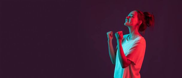 Portret pięknej młodej dziewczyny odizolowanej na ciemnym fioletowym tle studia w czerwonym świetle neonowym, filtr. Pojęcie ludzkich emocji, wyraz twarzy. - Zdjęcie, obraz