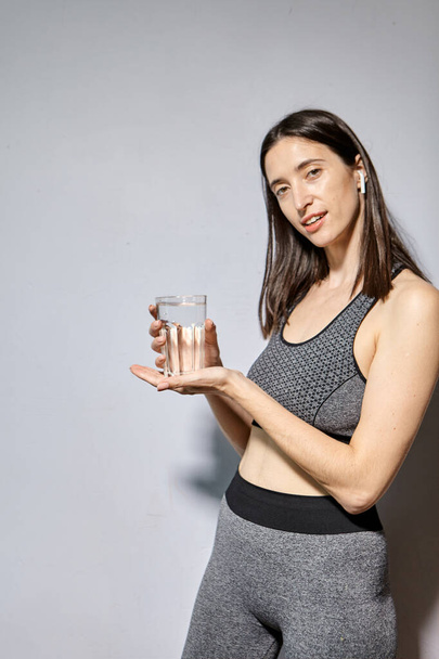 Μια γυναίκα με αθλητικά ρούχα και ασύρματα ακουστικά πίνει νερό. την έννοια του υγιεινού τρόπου ζωής και των αθλητικών δραστηριοτήτων - Φωτογραφία, εικόνα