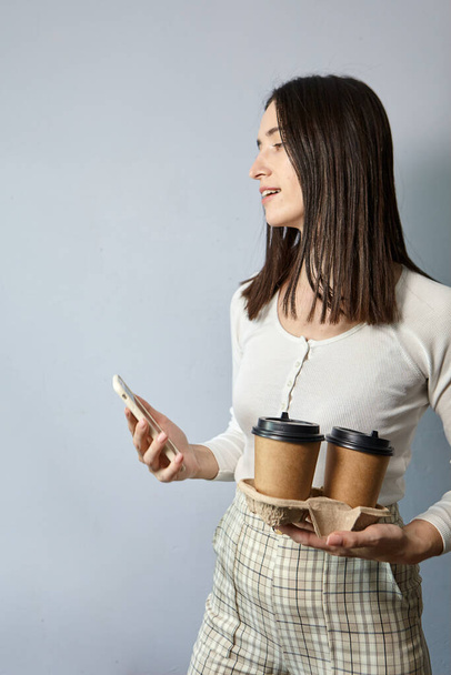 Una mujer con una sudadera blanca sostiene dos tazas de café en una mano, y un teléfono inteligente en la otra. Té o café para llevar. Comunicación remota - Foto, Imagen