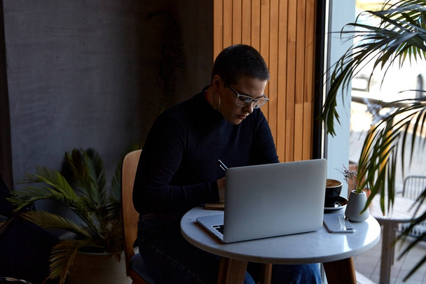 Egy fiatal lány szemüveggel, rövid hajjal és orrpiercinggel ül egy kávézóban és egy laptopon dolgozik. A szabadúszó és távoli munkavégzés vagy képzés fogalma. - Fotó, kép
