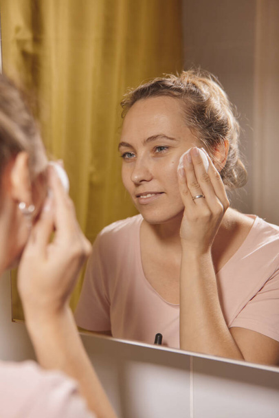 Een vrouw die haar gezicht schoonmaakt met een wattenschijfje in de badkamer thuis. vrouw in badkamer kijkt in spiegel en veegt make-up van haar gezicht met katoen pad. - Foto, afbeelding