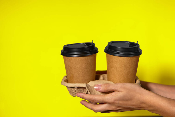 Κοντινό πλάνο σε κίτρινο φόντο, τα χέρια των γυναικών κρατούν δύο χάρτινα φλιτζάνια με καφέ σε ένα περίπτερο. Καφέ ή τσάι μαζί σου. Προώθηση δύο στην τιμή του ενός, παράδοση καφέ σε κίτρινο φόντο - Φωτογραφία, εικόνα