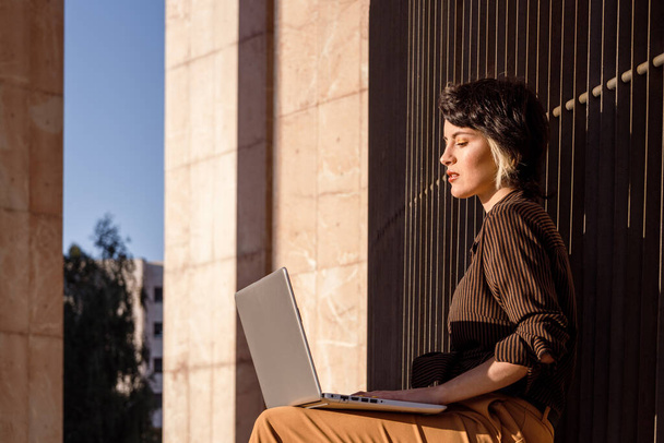 Młoda kobieta z krótkimi, nikczemnymi włosami i jesiennymi ubraniami pracująca w laptopie na świeżym powietrzu - Zdjęcie, obraz