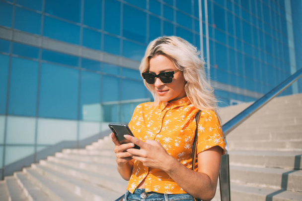 Beautiful female blogger in trendy sunglasses typing text for sending mobile message during online chatting at urban setting,youthful hipster girl connecting to 4g wireless internet for making booking - Zdjęcie, obraz