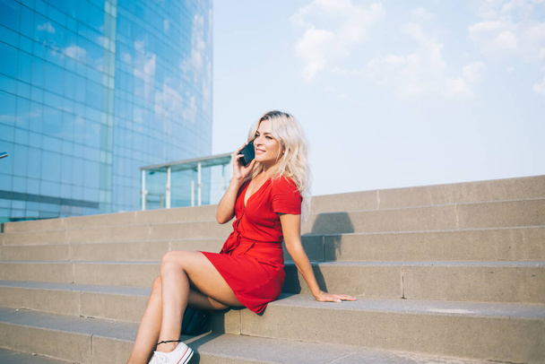 Carefree woman in stylish red sundress enjoying international contact conversation while resting at stairs, blonde female tourist receiving roaming phone call during summer vacations in urban city - Φωτογραφία, εικόνα