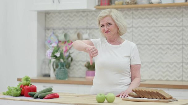 Стара жінка показує великі пальці вниз, стоячи на кухні
 - Фото, зображення