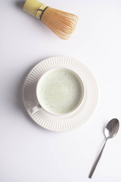 Een witte porseleinen beker met Japanse matcha thee drinken op een witte schotel op een wit oppervlak, een theelepel met matcha poeder en bamboe snor, bovenaanzicht - Foto, afbeelding