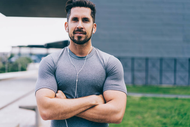 Half length portrait of handsome male athlete with muscular figure listening music during workout outdoors, handsome confident sportsmal looking at camera feeling determined reaching fitness goals - Photo, Image