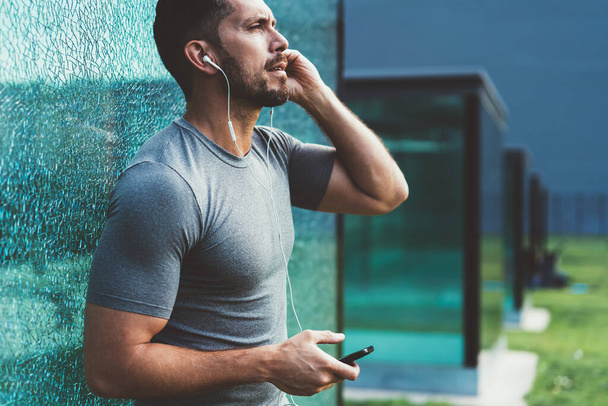 Confident handsome male athlete in activewear putting on earphones listening music via mobile phone application on street, serious sportsman using stereo equipment and smartphone for enjoy radio - Zdjęcie, obraz