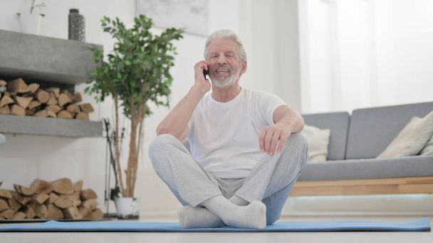 Alter Mann spricht zu Hause auf Fitnessmatte mit Smartphone - Foto, Bild
