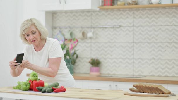 Stara kobieta robi zdjęcia owoców na smartfonie w kuchni  - Zdjęcie, obraz