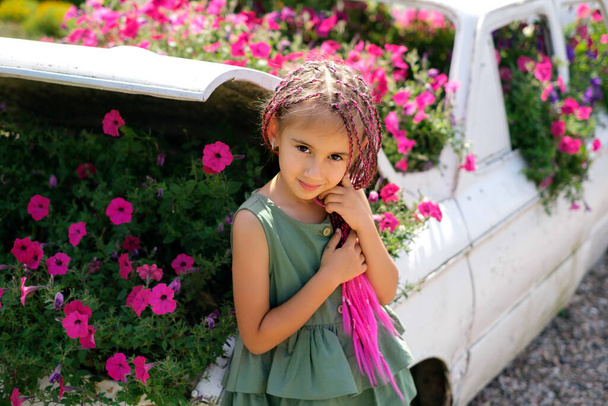 Una ragazza in un vestito con le trecce rosa degli zizi di kanekalon si trova in una macchina decorativa con fiori che crescono all'interno. Un bambino in un'aiuola con dei fiori. Un bambino moderno con un'acconciatura hippie - Foto, immagini