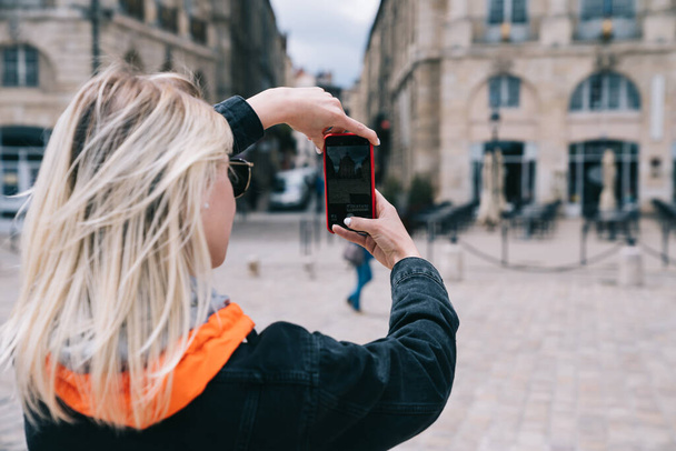 Back view of young female tourist standing on square in front of old buildings and taking photo with smartphone while walking and sightseeing in city - 写真・画像