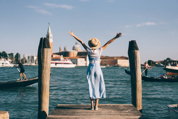 Back view of slim female in dress and hat stretching out arms and enjoying sunny day while standing on pier near canal in Venice, Italy - Фото, изображение