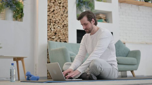Hombre joven usando el ordenador portátil en la estera de ejercicios en casa - Foto, imagen