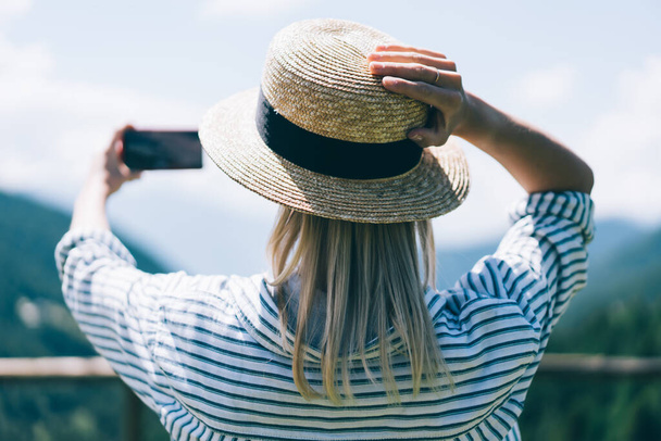 Back view of female traveler touching hat and using smartphone to take selfie while standing on observation deck during trip to Lago di Braies - Zdjęcie, obraz
