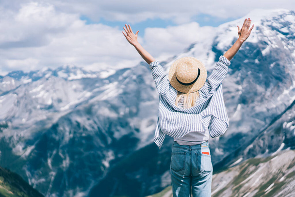 Back view of female traveler in hat raising arms and enjoying freedom while standing against mountain ridge during trip to Lago di Braies - 写真・画像