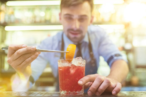 Ο μπάρμαν φτιάχνει ένα κόκκινο κοκτέιλ με ένα πορτοκάλι στο μπαρ. - Φωτογραφία, εικόνα