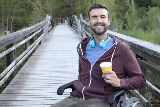 πορτρέτο του όμορφος νεαρός άνδρας σε αναπηρική καρέκλα κρατώντας χάρτινο φλιτζάνι καφέ σε ξύλινη γέφυρα στο πάρκο - Φωτογραφία, εικόνα