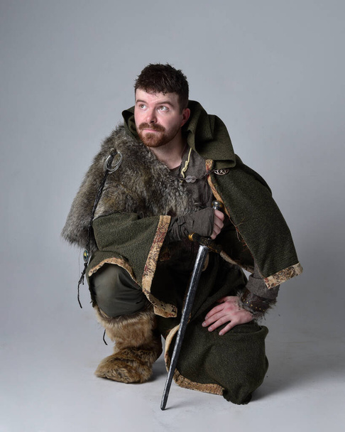 Teljes hossz portré fiatal jóképű férfi visel középkori kelta kalandor jelmez kapucnis köpeny, holding kard, elszigetelt stúdió háttér. - Fotó, kép