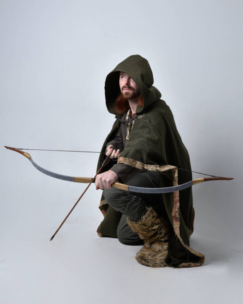 Teljes hossz portré fiatal jóképű férfi visel középkori kelta kalandor jelmez kapucnis köpeny, gazdaság íjászat íj és nyíl, elszigetelt stúdió háttér. - Fotó, kép