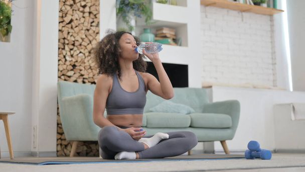 Mujer africana cansada bebiendo agua mientras hace yoga en casa - Foto, imagen