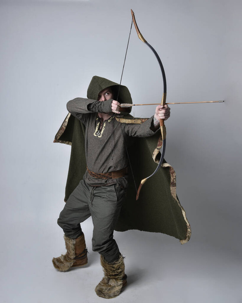 中世のケルトの冒険家の衣装を身に着けている若いハンサムな男の完全な長さの肖像画フード付きのマント,弓と矢を保持,スタジオの背景に孤立. - 写真・画像