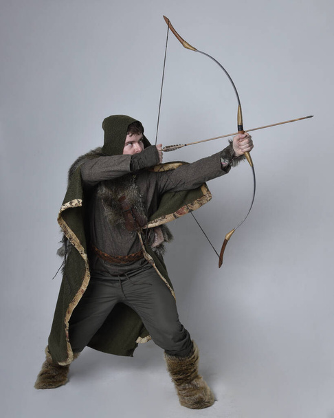 Retrato de comprimento total de homem bonito jovem vestindo traje aventureiro celta medieval com capa com capuz, segurando arco e flecha arco, isolado no fundo do estúdio. - Foto, Imagem