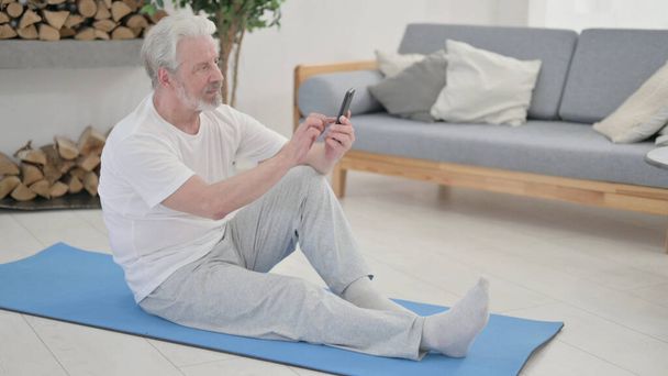 Oude man met Smartphone op Excercise Mat thuis - Foto, afbeelding
