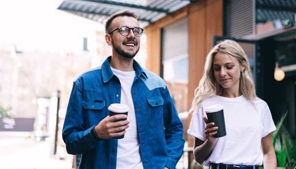 Positive young caucasian hipster couple in love holding coffee cup talking to each other strolling on street, smiling teen woman and man friends in casual wear satisfied with free time and dating - Foto, Imagem