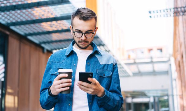 Pensive caucasian male checking banking notification on smartphone about online payment for coffee to go in cafe, serious hipster guy 20s holding beverage sending and texting messages on cellular - Foto, Imagem