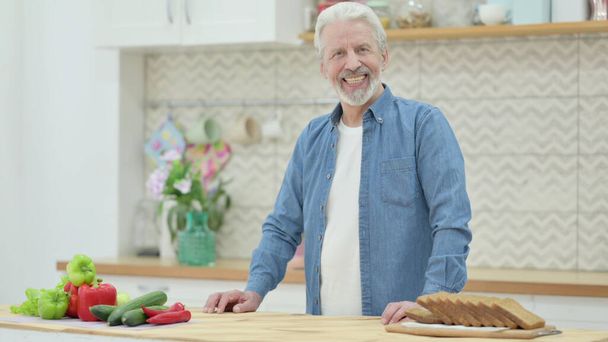 Alter Mann lächelt in die Kamera, während er in der Küche steht - Foto, Bild