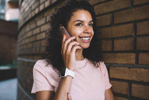 Glückliches dunkelhäutiges Hipstermädchen, das während eines positiven, freundlichen Smartphone-Gesprächs lächelt, fröhliche afroamerikanische Teenagerin, die die freie Zeit für die Kommunikation per Handy-Anwendung genießt - Foto, Bild