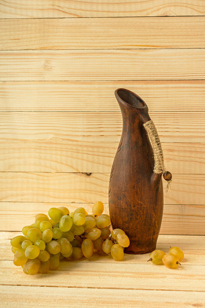 Grape jug on a wooden natural background. Rustic retro style. Antique clay jug with wine. Green juicy ripe grapes of the autumn harvest. the concept of agriculture, winemaking. Vertical still life - Photo, Image