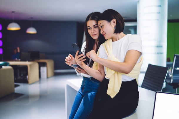 Pretty young multiracial female colleagues browse web page on smartphone reading news together during break, 20s women looking at common photo on smartphone for share in social networks blog - Fotó, kép