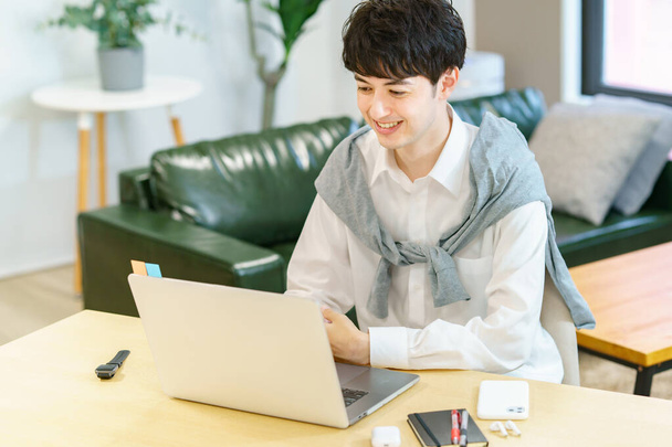 Asyalı genç bir adam sıradan bir odada dizüstü bilgisayar kullanıyor. - Fotoğraf, Görsel