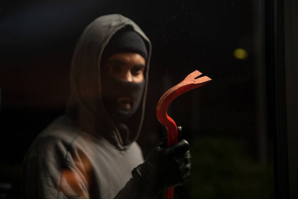 Burglar in balaclava with crowbar. Masked thief holding a red color lever, weapon and tool for vandalism and house break. Hooded hooligan in dark night background - Photo, Image