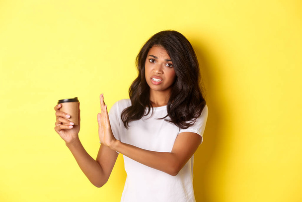 Afbeelding van Afrikaans-Amerikaans meisje klagen over slechte smaak van koffie, tonen afwijzing teken en weg te trekken kopje, staande over gele achtergrond - Foto, afbeelding