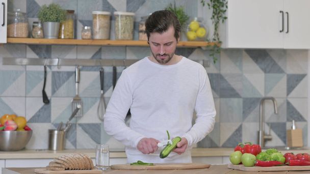Attractive Young Man Peeling Cucumber in Kitchen - Foto, immagini