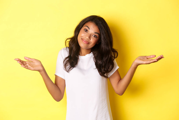 Portrait of clueless, beautiful african-american girl, shrugging and smiling, apologizing for being unaware, standing over yellow background - Foto, Imagen