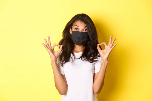 Concepto de coronavirus, pandemia y estilo de vida. Retrato de niña afroamericana complacida en máscara facial negra, mostrando signos de aprobación, como algo bueno, fondo amarillo - Foto, Imagen