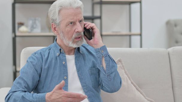 Старик разговаривает по смартфону, сидя на диване  - Фото, изображение