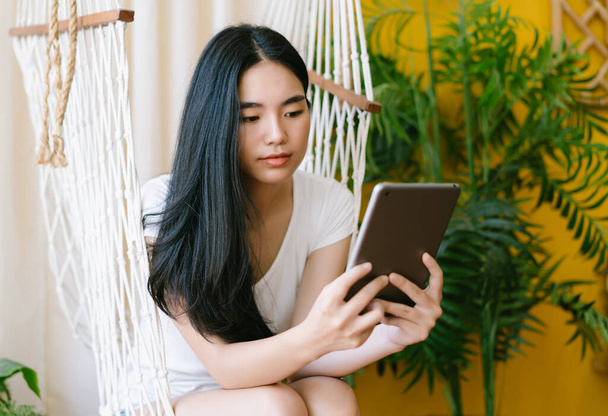 Portrét mladé asijské ženy, jak čte zprávu, e-book nebo informace na svém tabletu, zatímco sedí na houpačce v obývacím pokoji. Sociální média čas, moderní technologie snadné a pohodlné použití koncept. - Fotografie, Obrázek