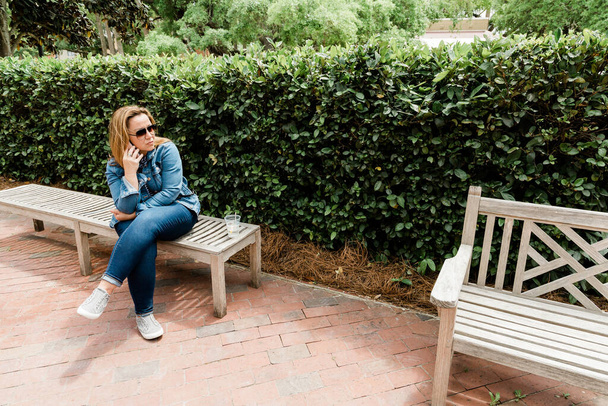 Μια γυναίκα με γυαλιά ηλίου που κάθεται σε ένα παγκάκι και μιλάει στο τηλέφωνο - Φωτογραφία, εικόνα