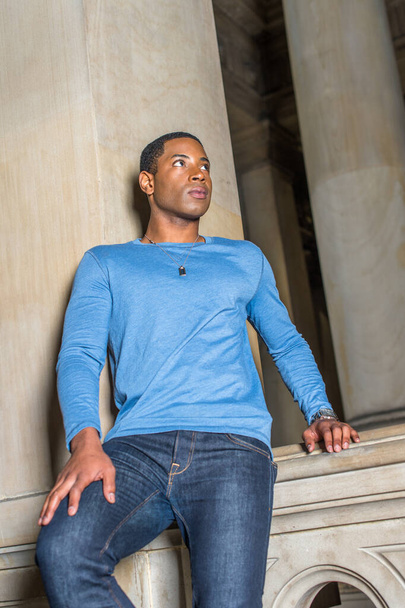 Habillé d'un T-shirt bleu clair à manches longues, un jeune étudiant noir séduisant se tient debout dans un couloir contre un mur, levant les yeux et réfléchissant - Photo, image