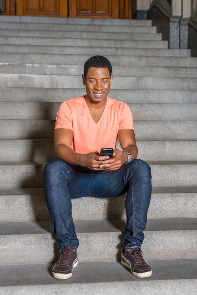 Одеваясь в светло-оранжевый короткий рукав V рубашка шеи, джинсы, молодой красивый черный студент сидит на лестнице возле офисного здания, проверяя сообщения на мобильном телефоне. - Фото, изображение
