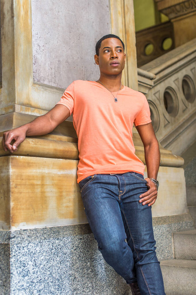 Vêtue d'une chemise à manches courtes en V orange clair, d'un jean, une jeune étudiante noire séduisante se tient debout en bas contre une colonne, impatiente - Photo, image